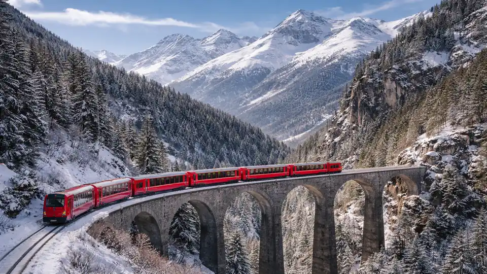 Bernina Express überquert ein historisches Viadukt vor verschneiten Alpen, umgeben von winterlichen Nadelwäldern und schroffen Berggipfeln im klaren Tageslicht.